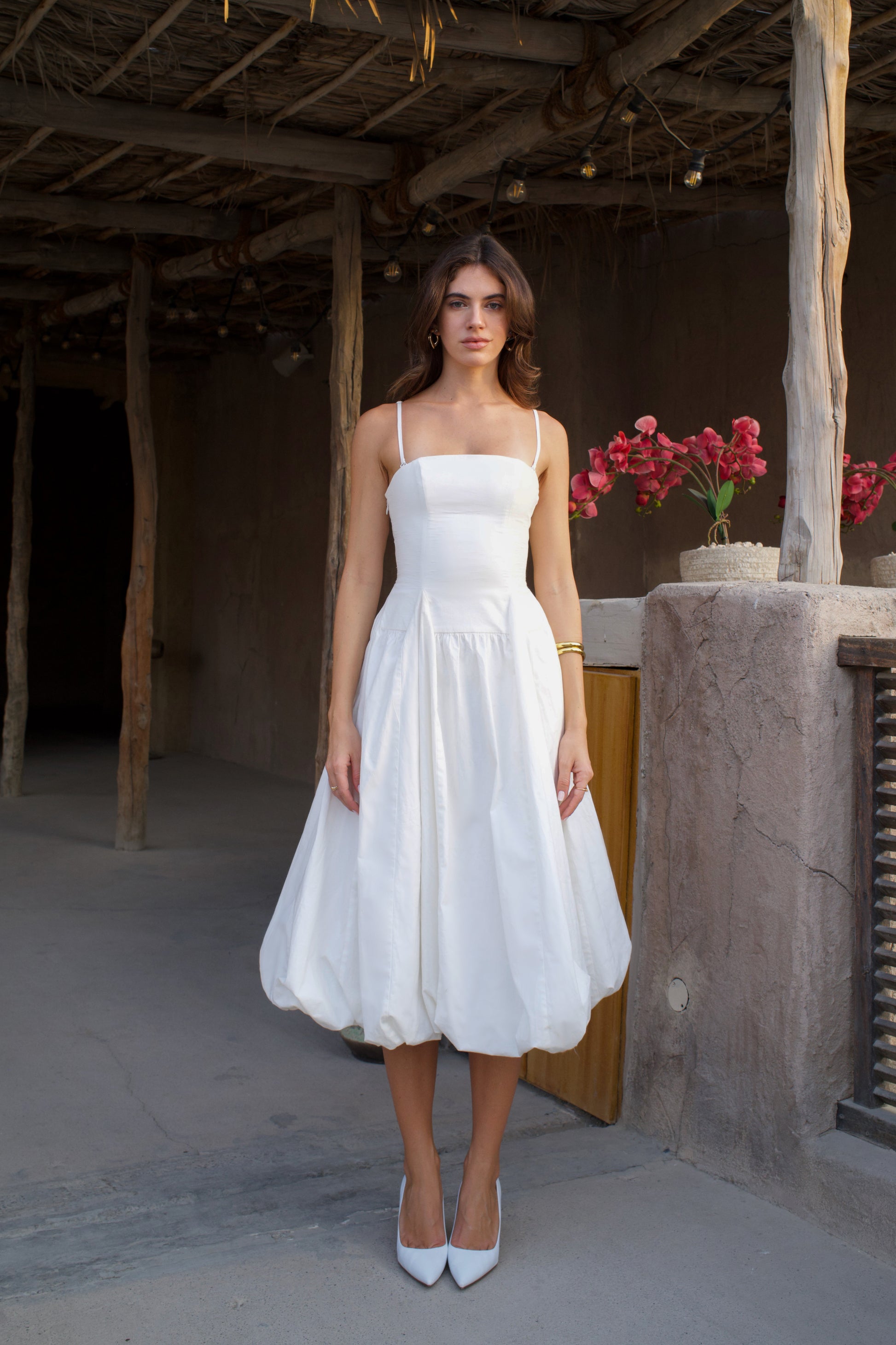 Woman in a white dress standing in a rustic outdoor setting with flowers in the background.