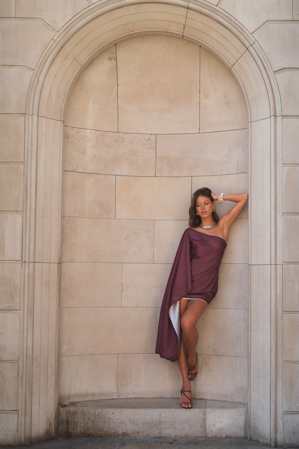 A woman posing in a draped plum purple one-shoulder dress against a beige stone archway background.