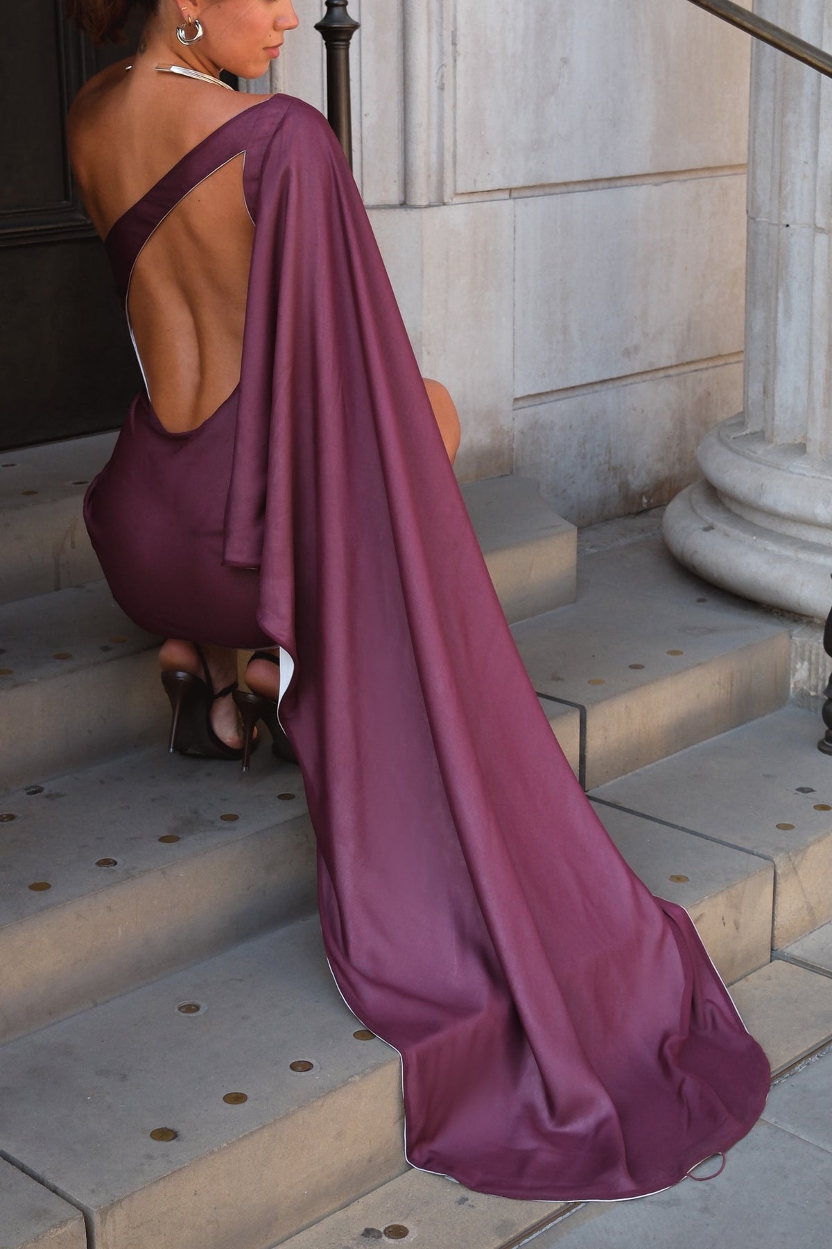 A woman wearing a plum purple one-shoulder dress with a dramatic open back and long flowing train, sitting on stone steps in front of a classical building.