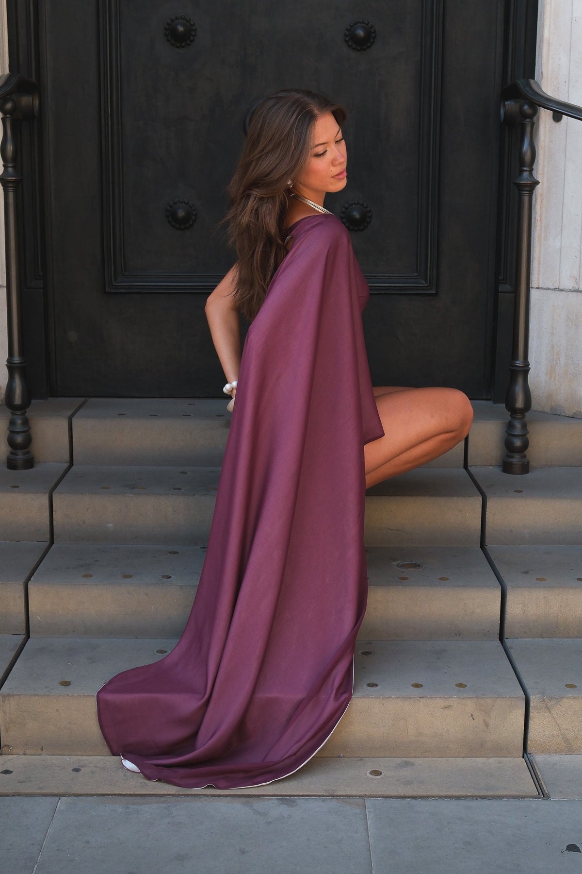 A woman wearing a flowing mauve-purple cape-style dress sits on stone steps in front of a dark wooden door.