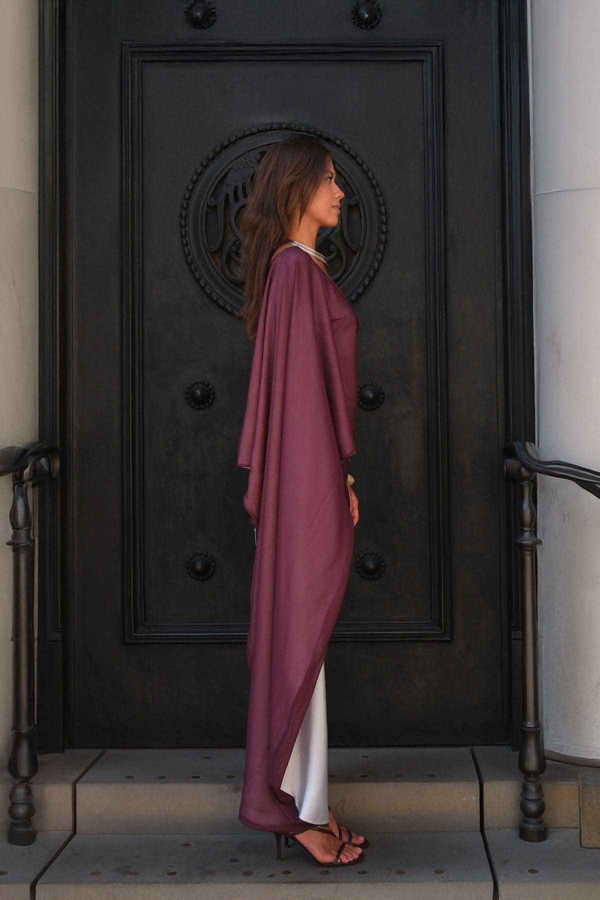 A woman stands in profile in front of a large black decorative door, wearing a flowing mauve-purple kaftan dress over white trousers and black sandals.