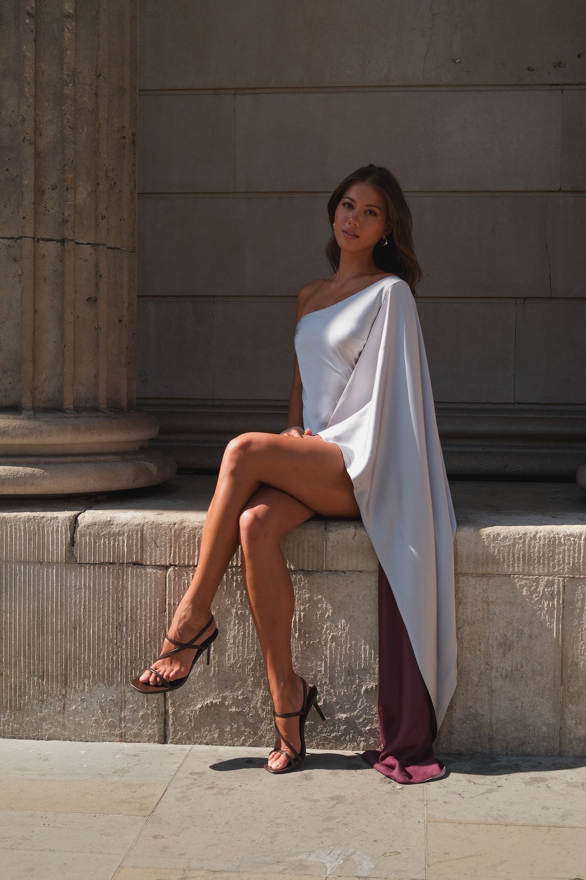 A woman sits on a stone ledge outdoors, wearing a one-shoulder draped white dress with a deep purple side panel and a small pink accent at the hem, paired with black strappy heels.