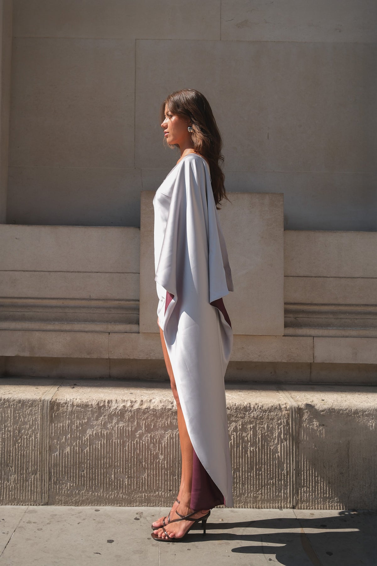 A woman stands in profile outdoors against a beige stone wall, wearing a flowing draped white dress with a high slit and dark strappy heels.