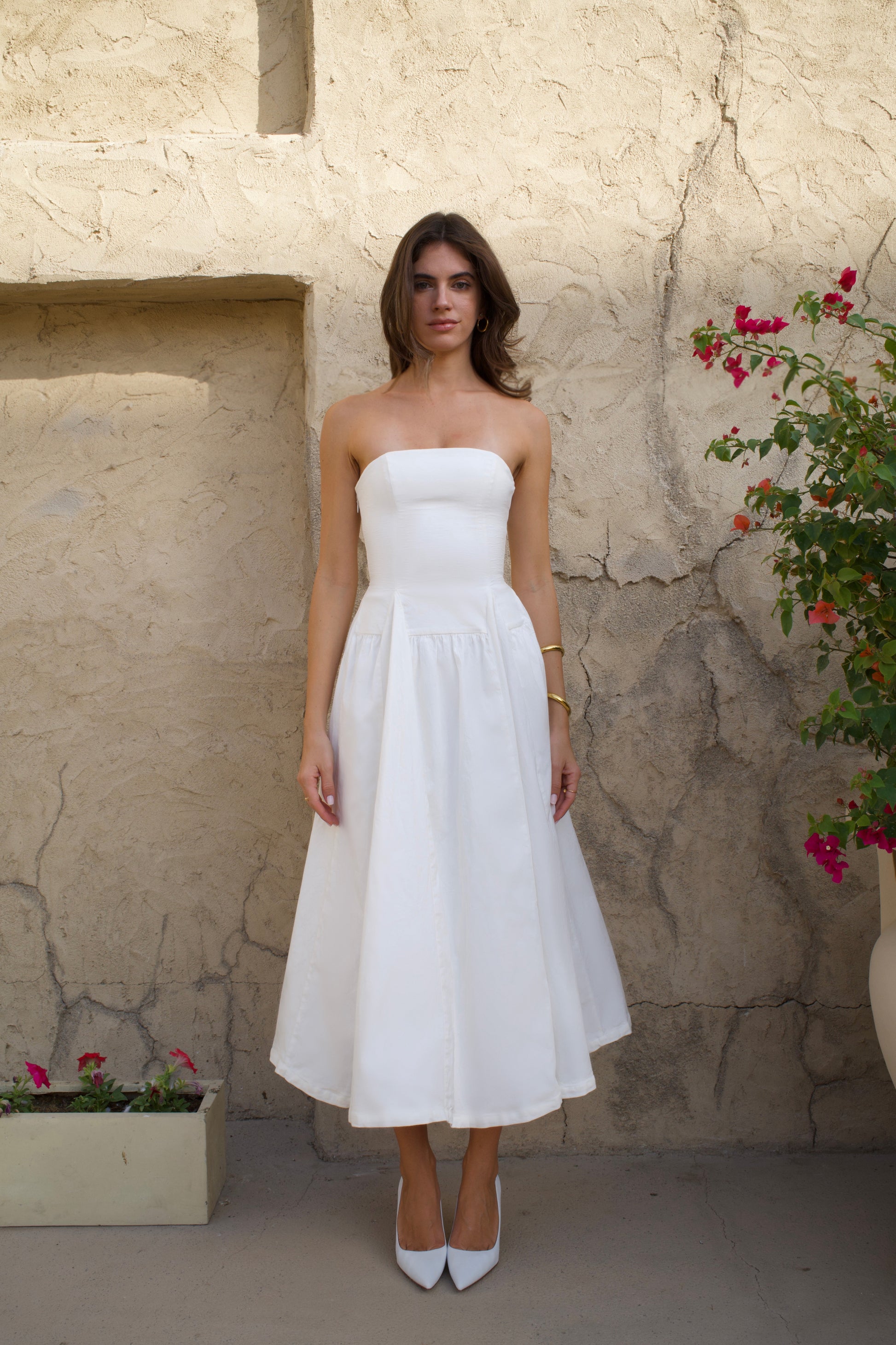 Woman in a white strapless dress standing against a textured stone wall with flowers.